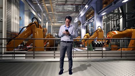 full body of an asian male professional worker standing with his smartphone with industrial robotic arms working, working continuously