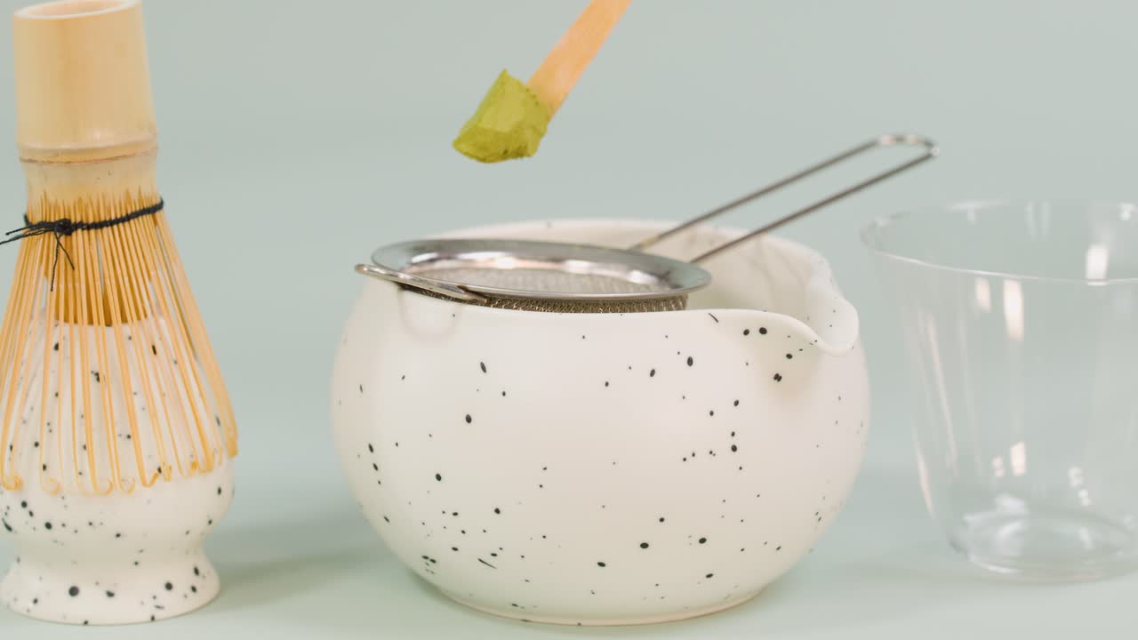 Hand sifts green matcha powder into bowl using bamboo spoon, soft lighting, static camera