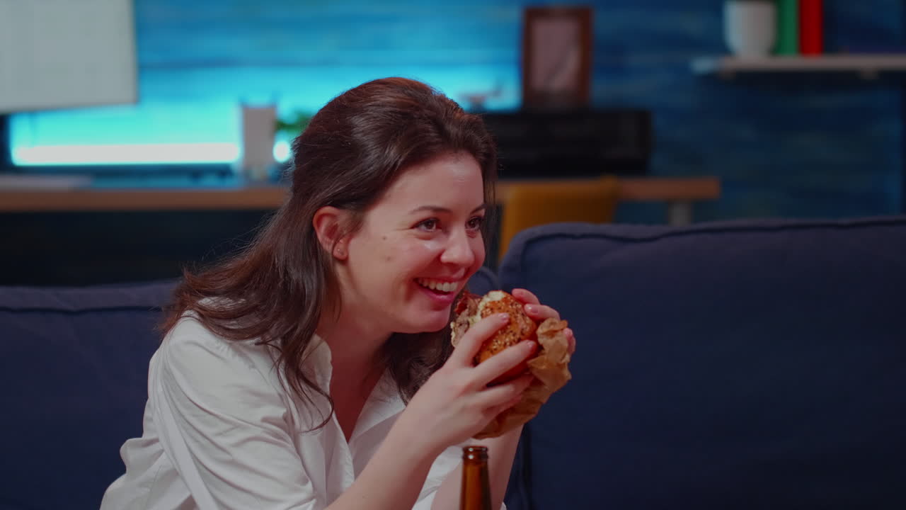 Young person laughing at TV while eating hamburger