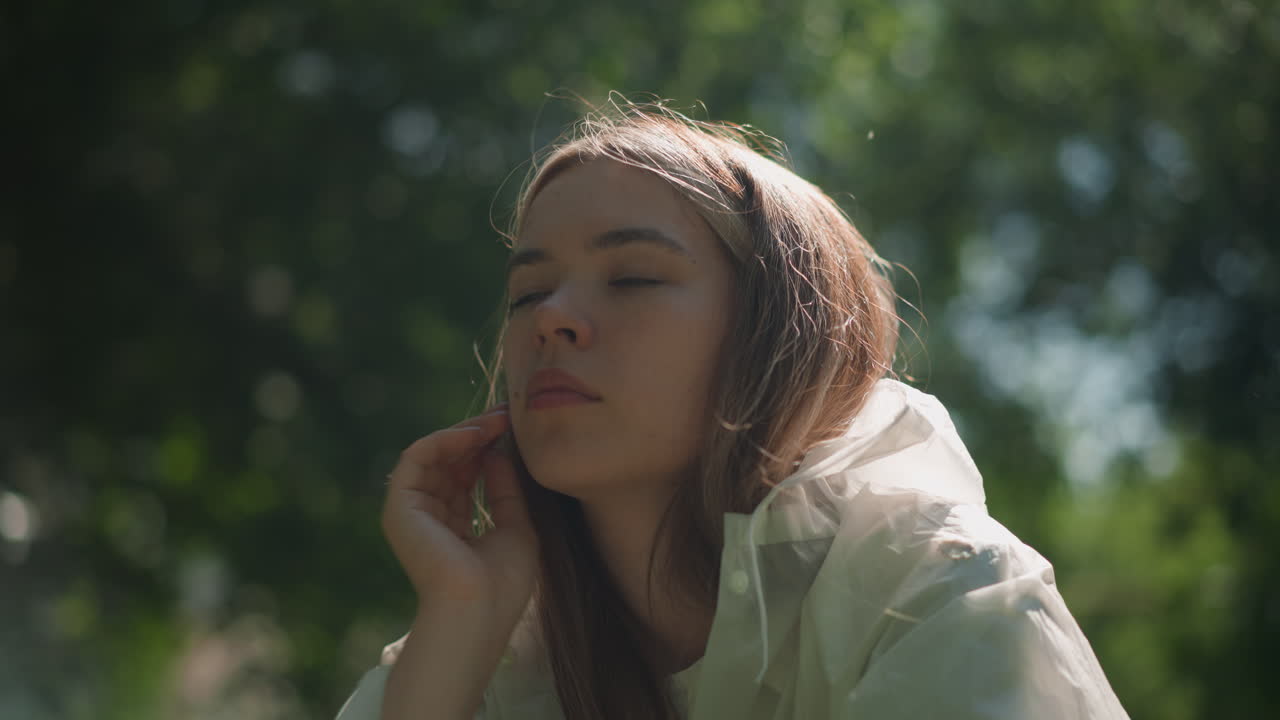 joven con impermeable translúcido baja su capucha, inclinándose hacia adelante pensativo como la luz del sol se refleja en su cara contra un fondo verde exuberante