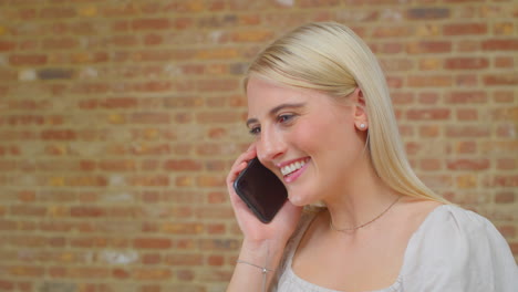 Mujer-Joven-Parada-Junto-A-Una-Pared-De-Ladrillos-En-Casa-Hablando-Con-Amigos-Por-Teléfono-Móvil