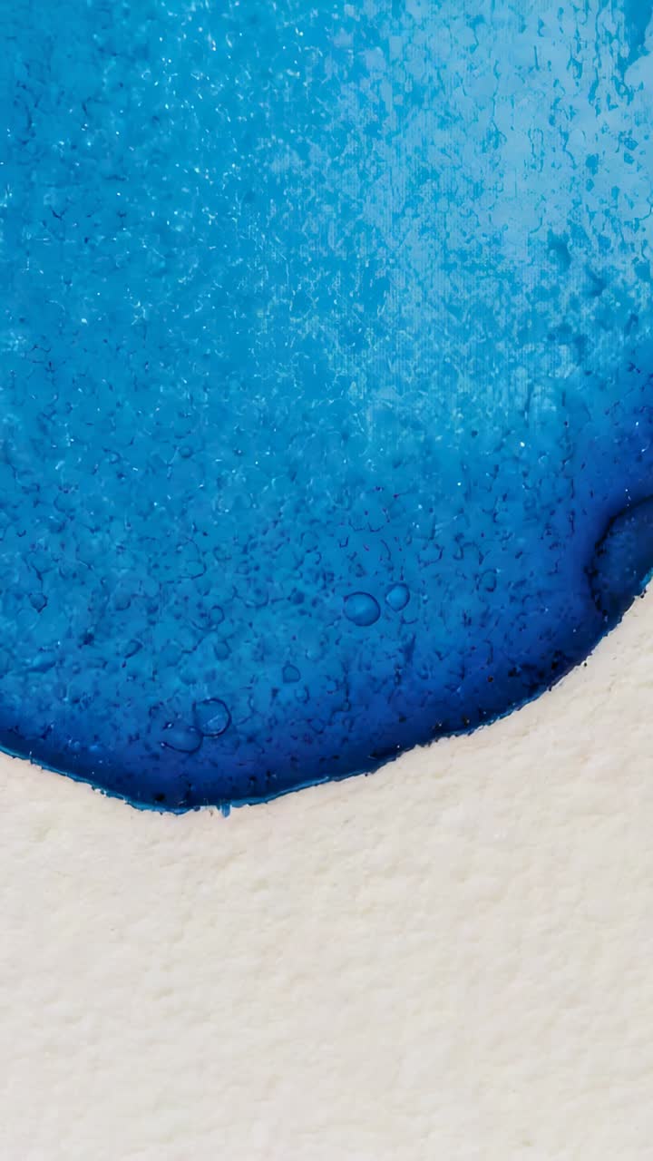 Vertical video: Evaporating blue wash settling on textured watercolor paper on table, with droplets