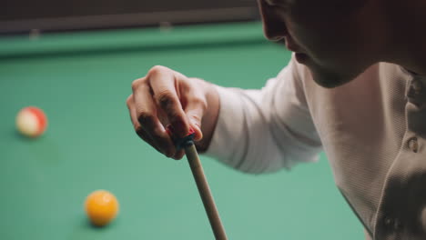 player in white shirt chalking cue stick before taking shot on green pool table. close-up highlights preparation, focus, and skill, with blurred billiard balls in background