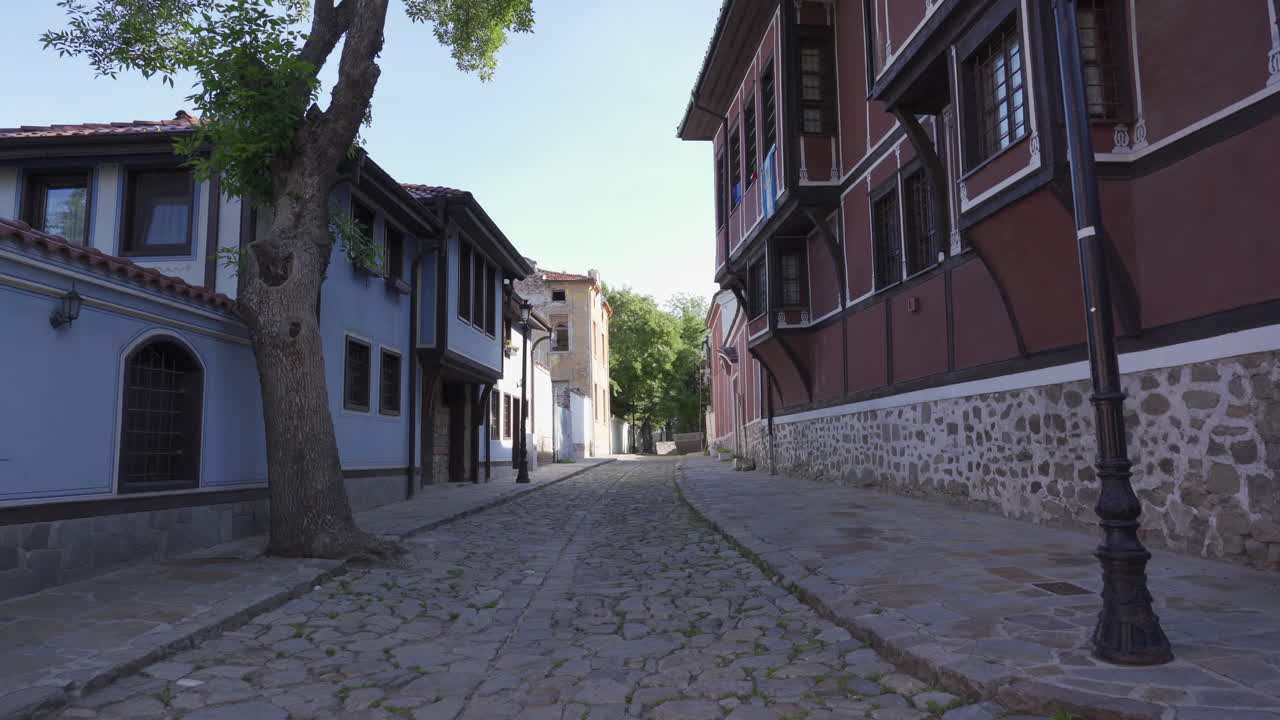 Carrellata lungo una tranquilla via acciottolata nel centro storico di Plovdiv, fiancheggiata da case in stile rinascimentale bulgaro e muri in pietra, filmata nella tenue luce del mattino