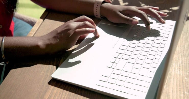 Hands are typing on light laptop at sunlit table as right hand is lowering for work