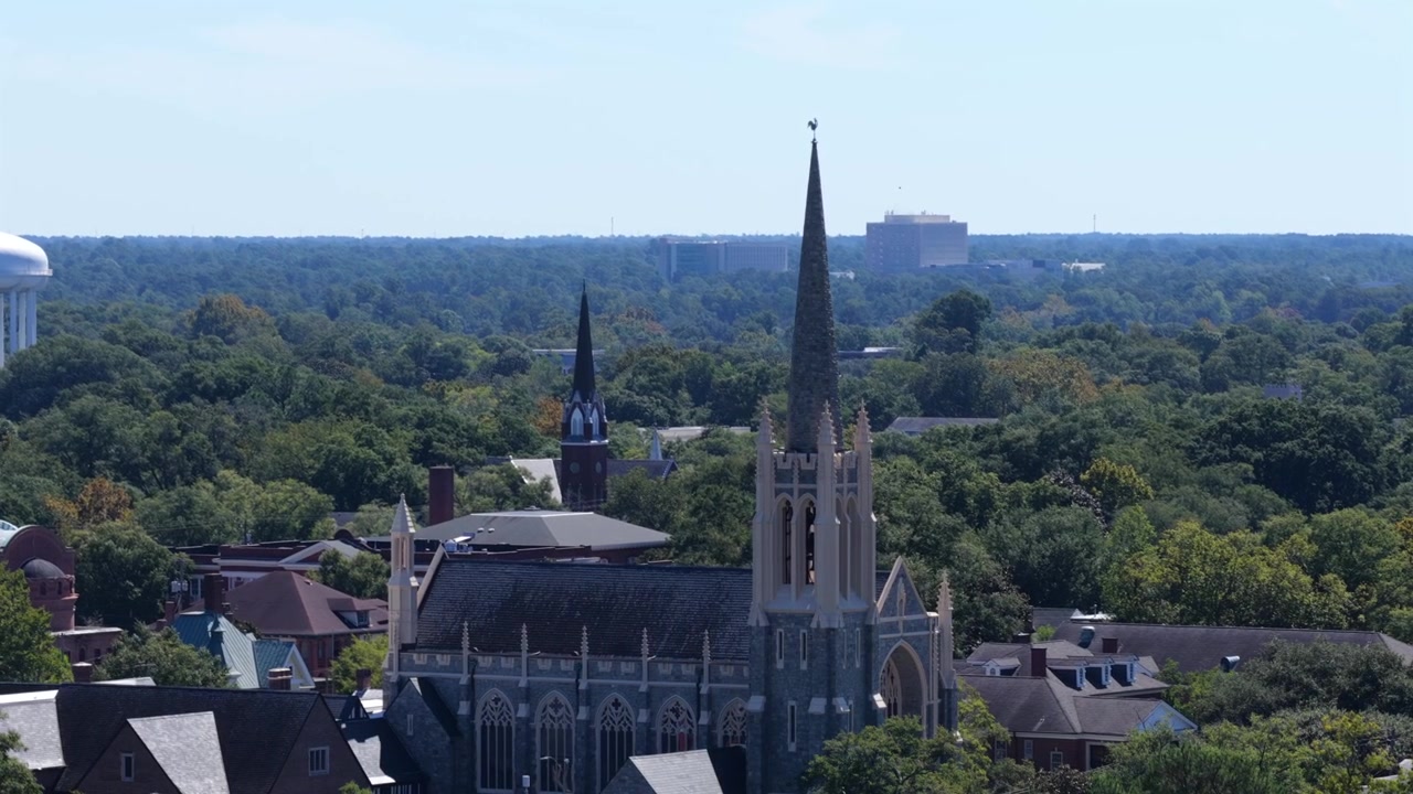 Uma torre de igreja gótica alta ergue-se acima de uma densa copa de árvores e telhados de ardósia em Wilmington, Carolina do Norte, com uma torre de água branca e um bloco de escritórios distante no horizonte. Vista aérea