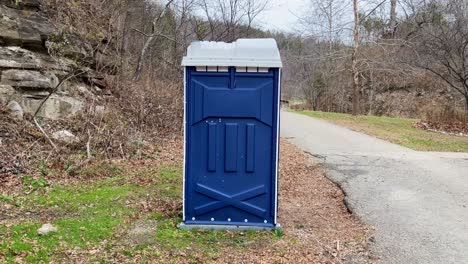 Video-Estático-De-Un-Orinal-Porta-Azul-En-Un-Parque-Con-árboles-Muertos-Y-Caminos-Asfaltados