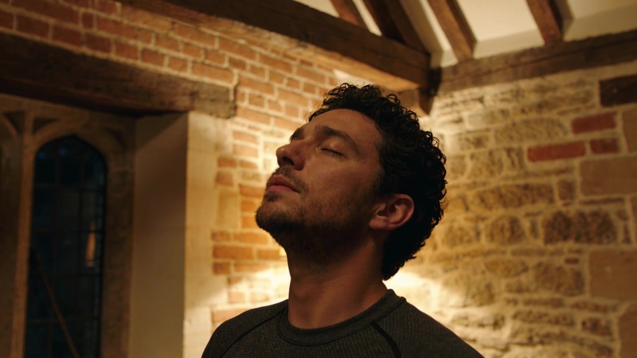 Un uomo con gli occhi chiusi, che reclina la testa all'indietro in un loft rustico, indossando un top scuro, rilassandosi accanto a un muro di pietra.