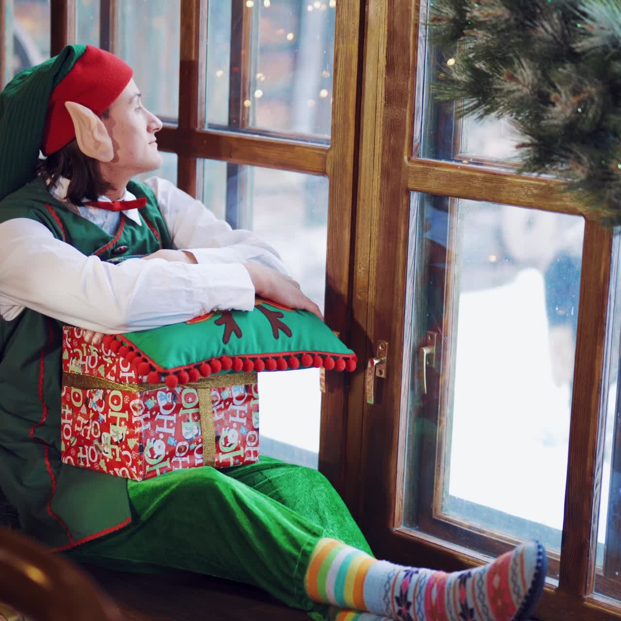 Elf in a green suit sitting on the windowsill with a gift in his hand and a New Year's pillow and looking out the window at the deer waiting for a miracle on the background of the Christmas tree in the room during the day