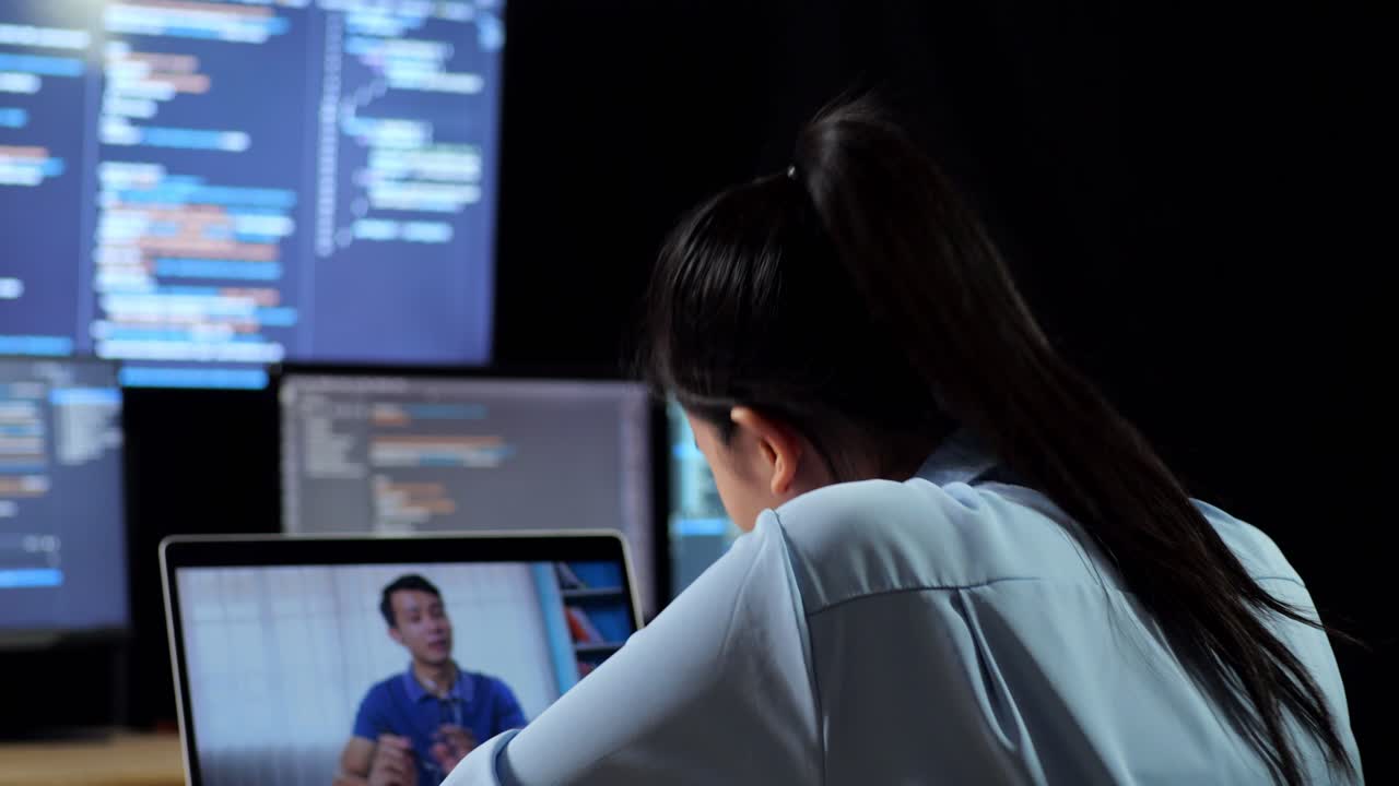 vista de atrás de una programadora asiática teniendo una reunión junto a una computadora portátil mientras escribe código en múltiples monitores que muestra la base de datos en los escritorios de ventana de terminal en la oficina