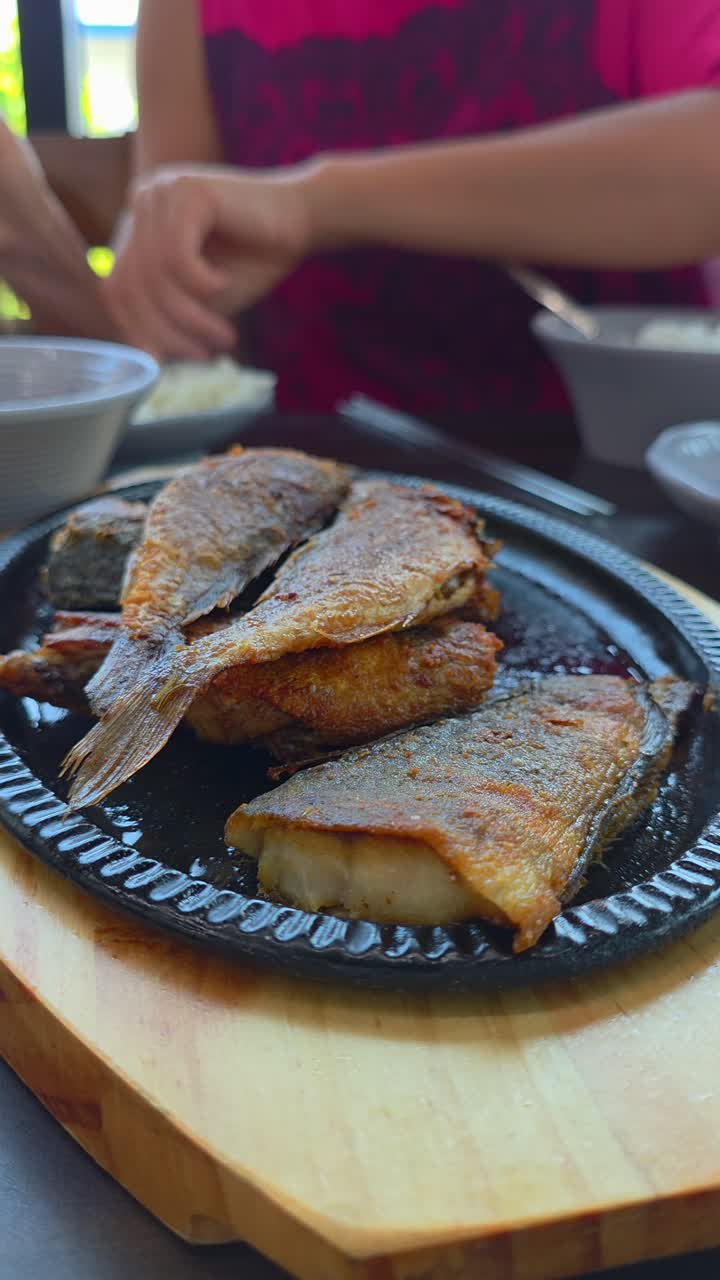 A close-up shot shows a woman using metal chopsticks to pick up a piece of traditional Korean grilled fish, saengseon-gui, served sizzling on a hot cast-iron plate at a restaurant in Korea - vertical
