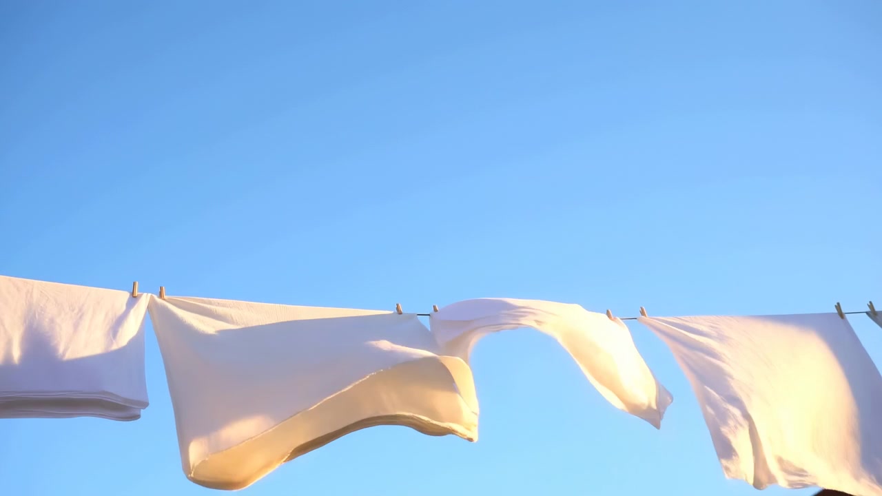 White sheets billowing on clothesline beneath clear blue sky, moving in warm golden sunlight and gentle breeze, creating calm domestic outdoor scene with soft shadows and airy fabric texture