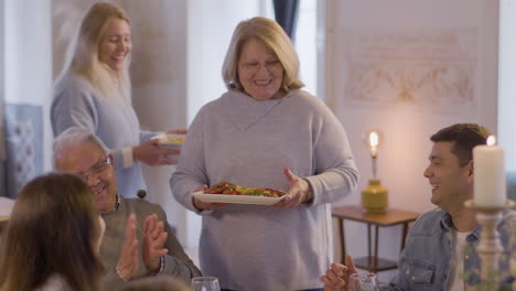 happy family sitting at table in dining room, then two women bringing plates with food and everyone cheering