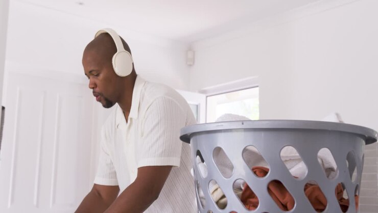 African man taking shirt from grey basket, folding and stacking clothes at counter with headphones