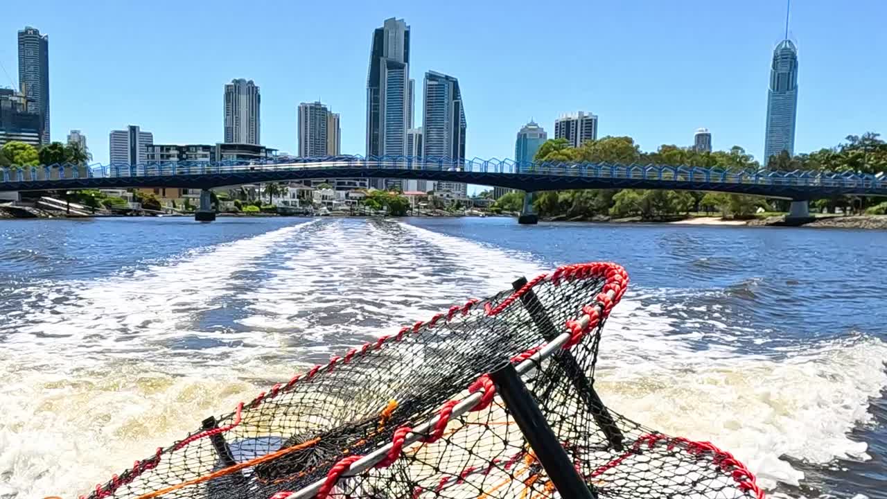 A crab pot on a boat travels along a river with a city skyline in the background.