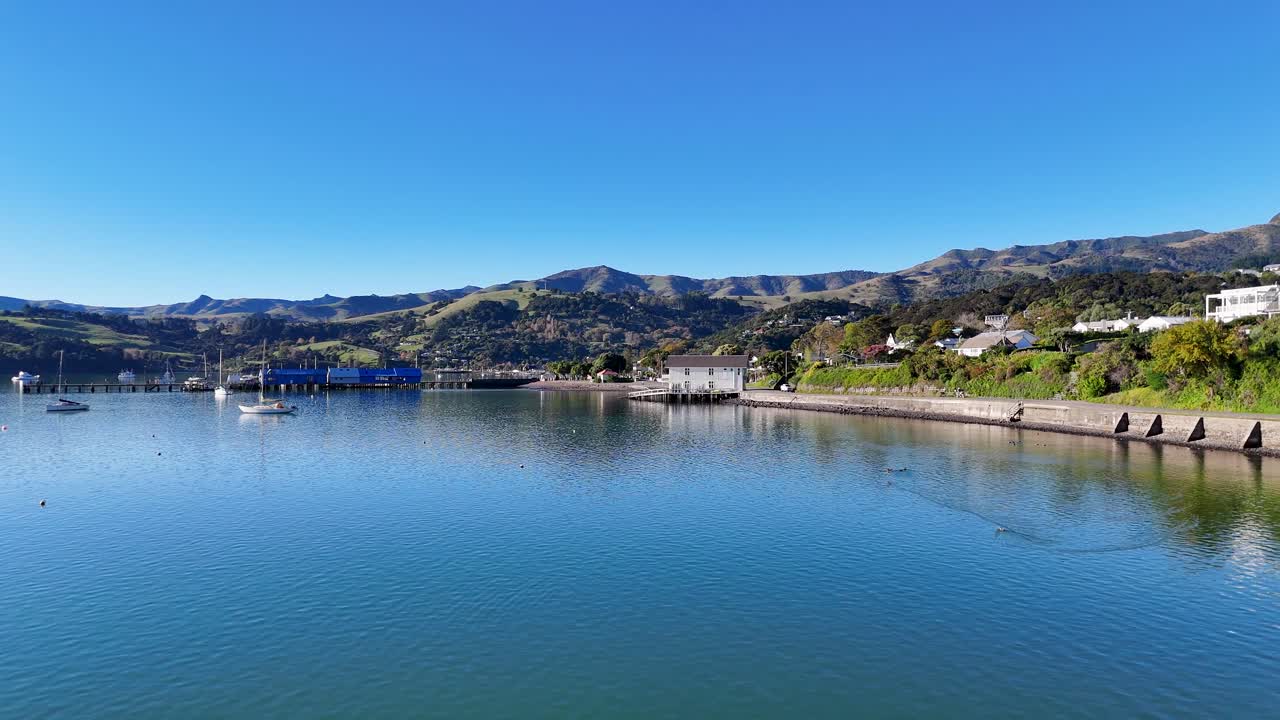 Aerial footage captures Akaroa's tranquil bay with calm waters, lush greenery, and distant hills under clear blue skies