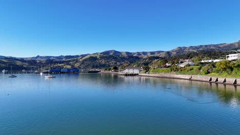 aerial footage captures akaroa's tranquil bay with calm waters, lush greenery, and distant hills under clear blue skies
