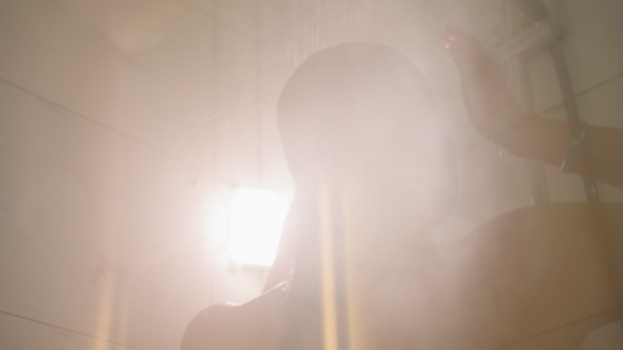 Hotelgast unter der Dusche spült sich die Haare unter warmem Wasser, sanfter Dampf und Gegenlicht erzeugen eine Silhouette, Glasabtrennung und Wassertropfen auf der Haut, ruhige Hotelbad-Stimmung, intimer Moment der Besinnung.