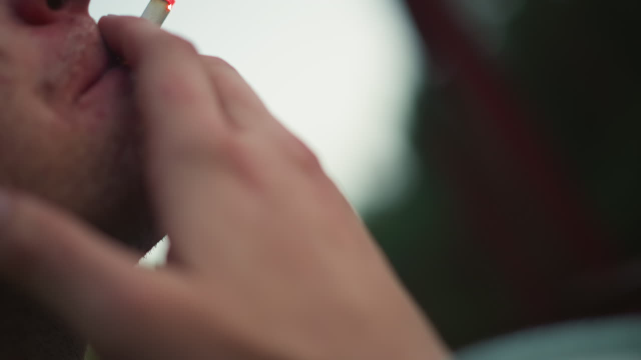 Uomo bianco che accende una sigaretta con le mani, primi piani delle dita che fanno scintille con l'accendino, brace che si illumina sulla punta della sigaretta, labbra che aspirano il fumo, rituale tattile catturato con profondità di campo ridotta, pausa a bordo strada