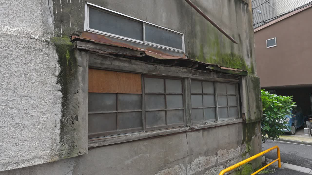 An old, weathered urban building in Japan features boarded-up windows and peeling walls covered in moss, suggesting a sense of decay and history.