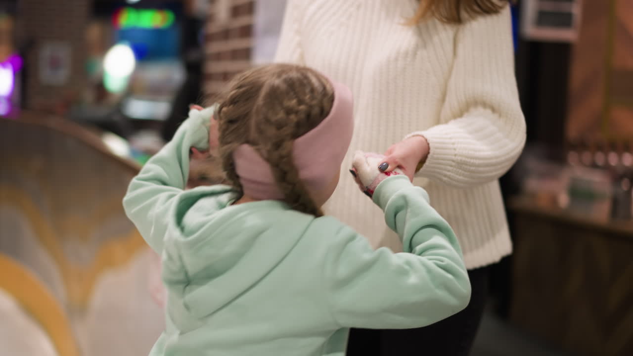 vista de cerca de una niña en una chaqueta verde menta y gorra rosa, extendiéndose para abrazar a un adulto en el borde de la pista de hielo