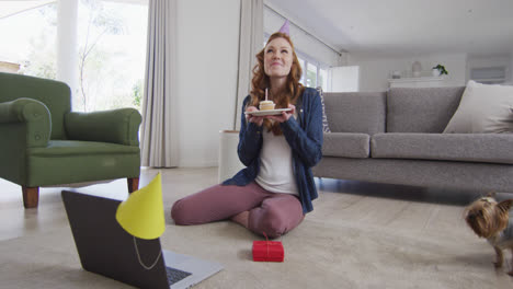 woman in party hat blowing candle on the cake while having a video chat on laptop at home