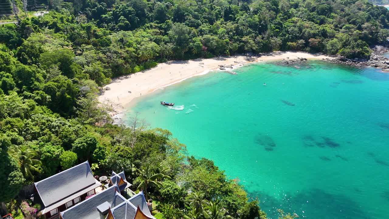Un filmato aereo cattura una spiaggia serena a Phuket, Thailandia, con acque turchesi e vegetazione lussureggiante sotto la luce del sole splendente.