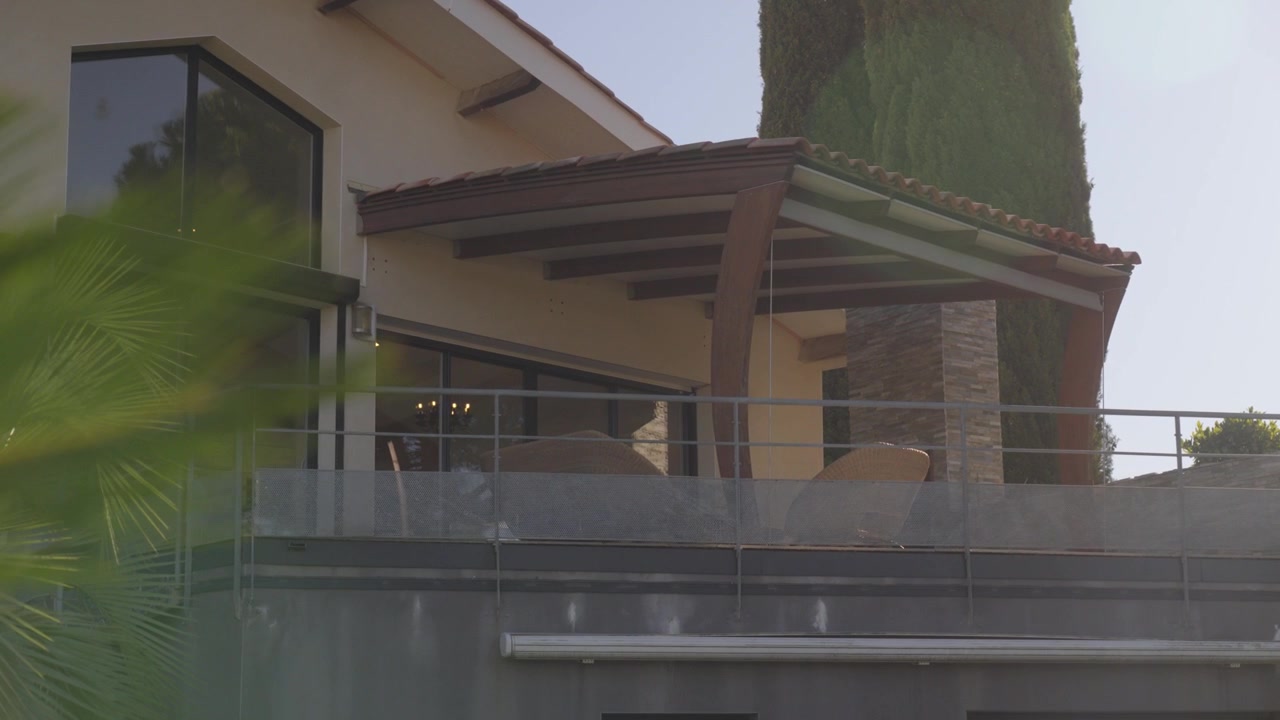 Una toma lenta detrás de una fronda de palma verde, revelando la terraza balcón de una hermosa casa al final del día