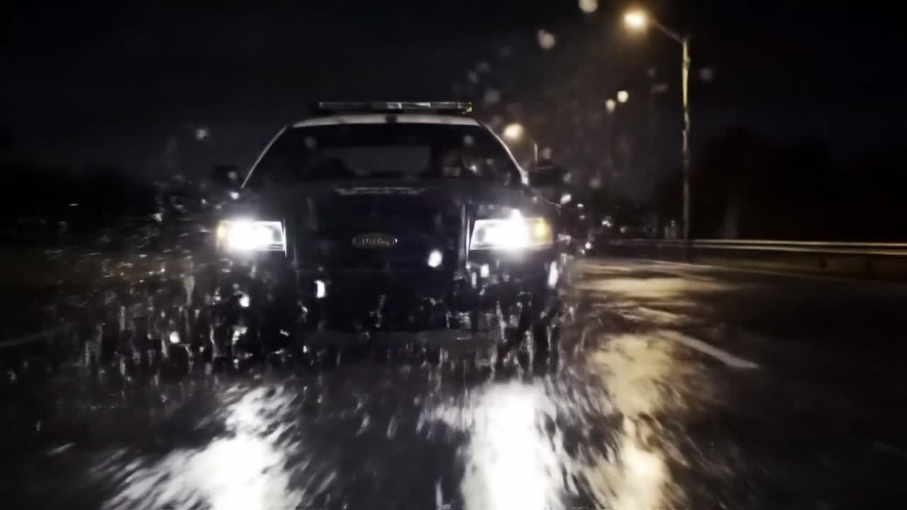 A high-speed pursuit unfolds on a wet highway at night, with a police vehicle closely trailing a fleeing car through puddles reflecting lights.
