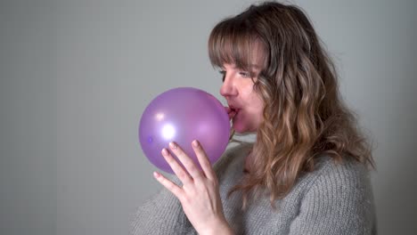 mujer joven que sopla un globo púrpura contra un fondo gris