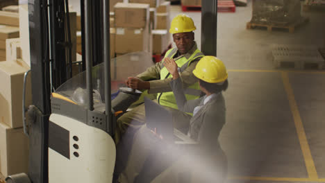 african american male and female workers wearing safety suits and using laptop in warehouse