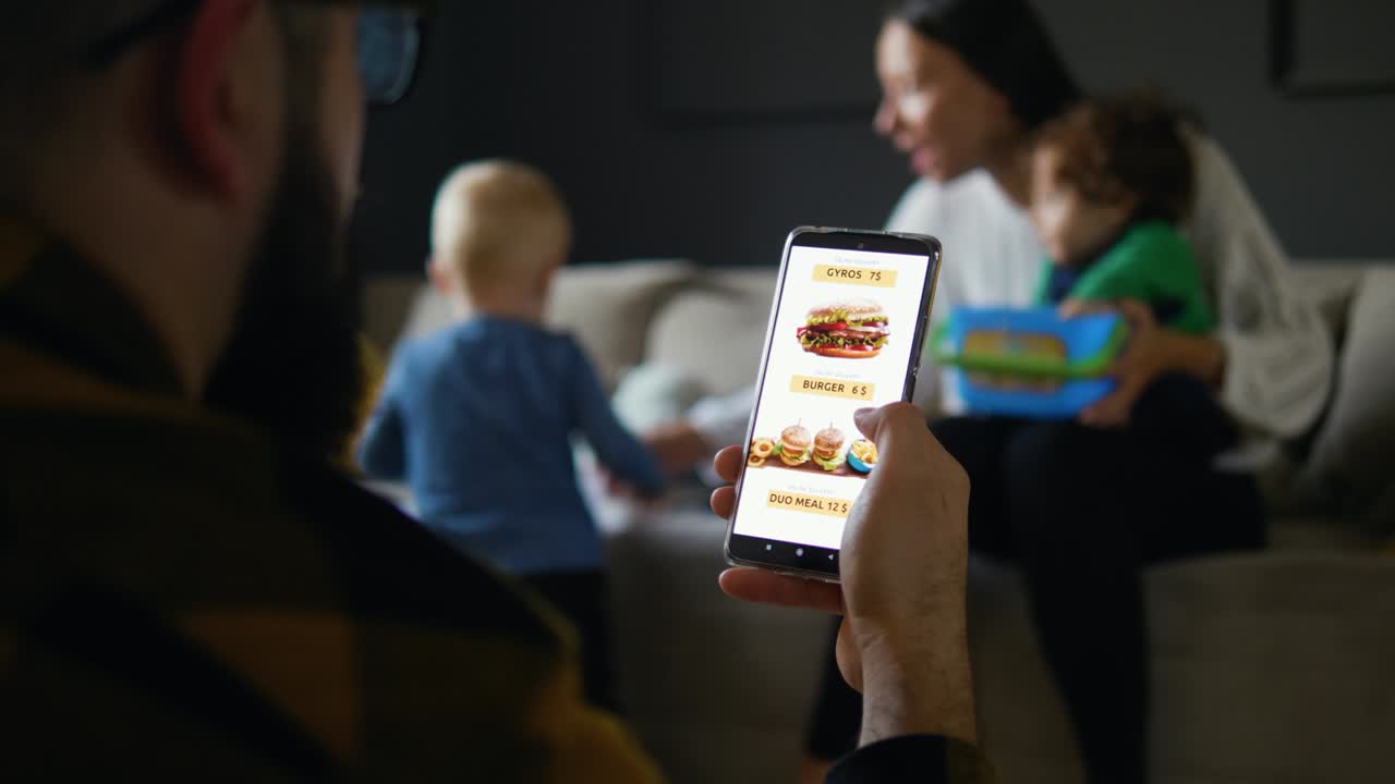 Man at Home sitting on the floor in Living Room Ordering Food Meal on mobile device app. Father orders food online using the phone while the young mother plays with the children in the background.