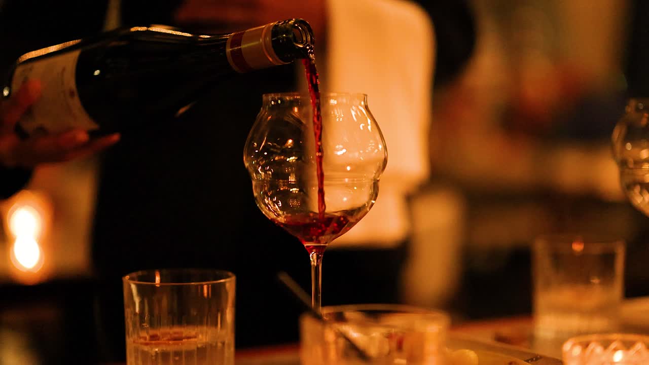 A bottle of red wine is poured into a glass in a warmly lit dining environment, creating a sophisticated and inviting atmosphere