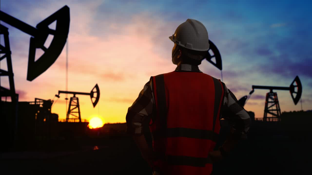 Back View Of A Male Engineer Wearing Safety Helmet Looking Around While Standing With Arms Akimbo In Front Of The Oil Pumps, During Sunset Or Sunrise Time