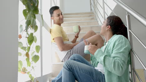 happy diverse gay male couple sitting on sunny staircase having coffee and talking, slow motion