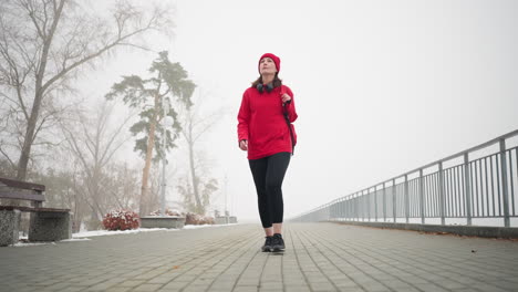 mujer de negocios con chaqueta roja de invierno y calentador de cabeza caminando por un camino pavimentado de polvo de nieve en una atmósfera de niebla, los auriculares descansan alrededor de su cuello, con árboles y bancos cerca