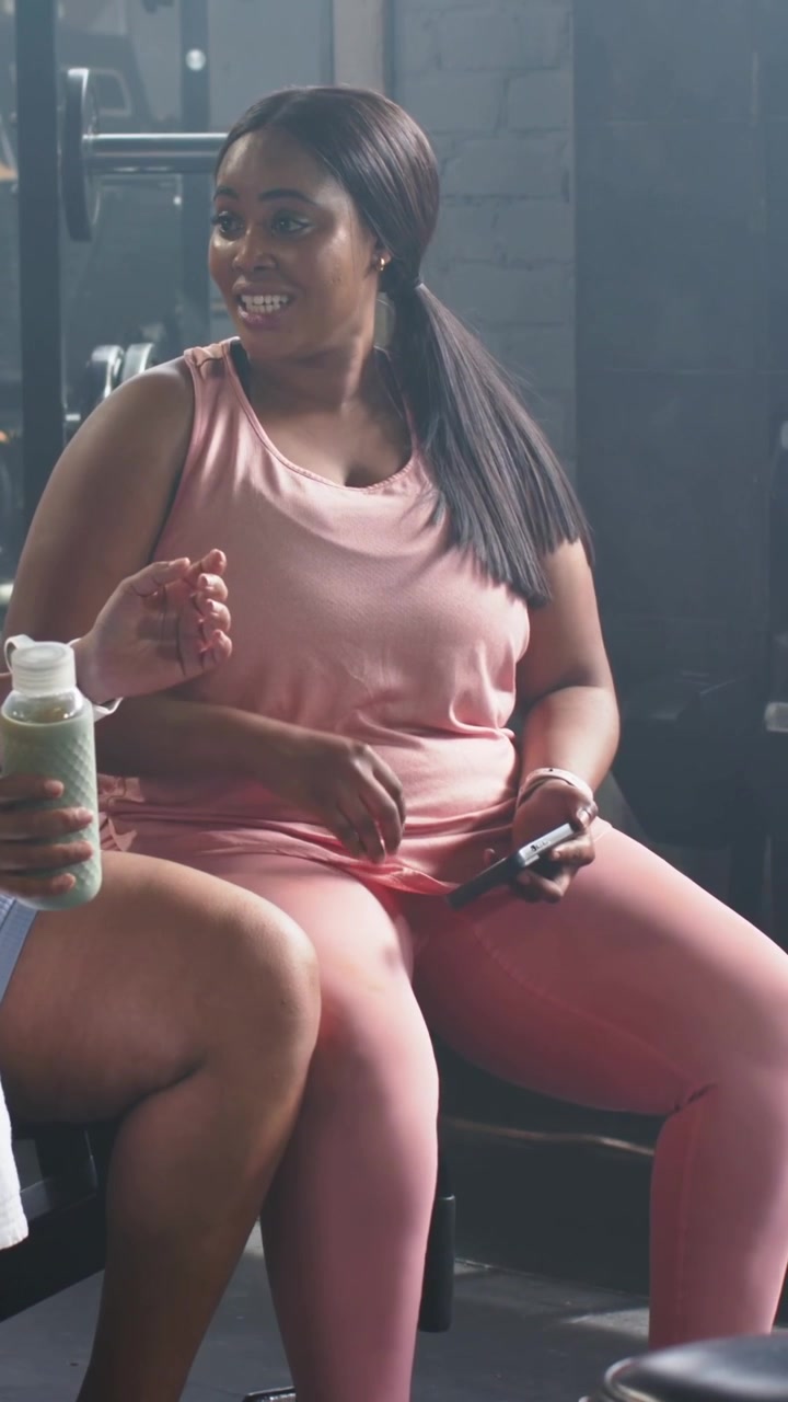 Video verticale: Donna in rosa che si gira e risponde a un'amica che parla in palestra, con il telefono vicino a una bottiglia verde.