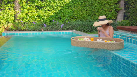 woman sitting in a swimming pool with floating tray of breakfast smelling coffee