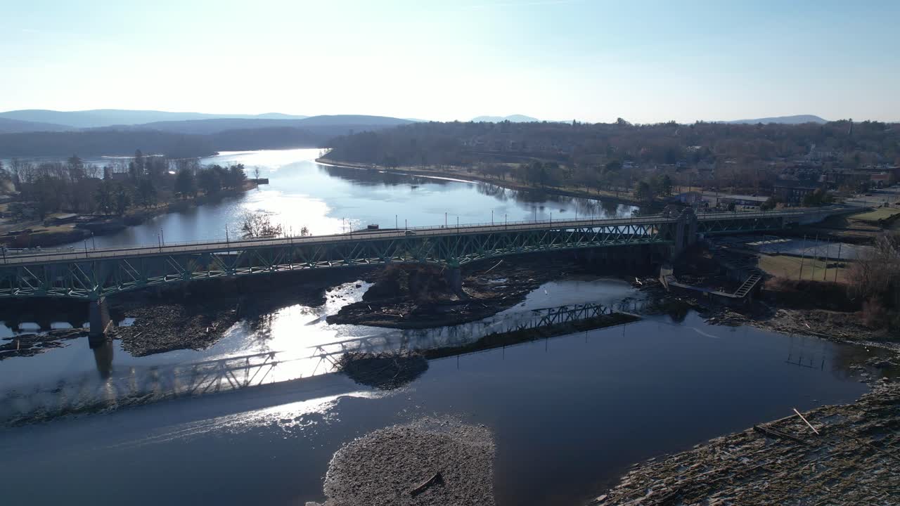 Side tracking along historic New England bridge over dams and waterways with early sunlight shimmering and a rocky riverbed in foreground