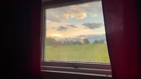 a countryside field with scattered trees and a house in the distance, seen through a window with red curtains under a cloudy sunset sky