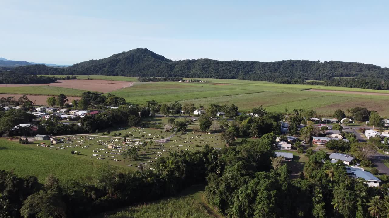 Drone ascending over a small country town showing the town cemetery and a large sugarcane plantation with mountains in the background