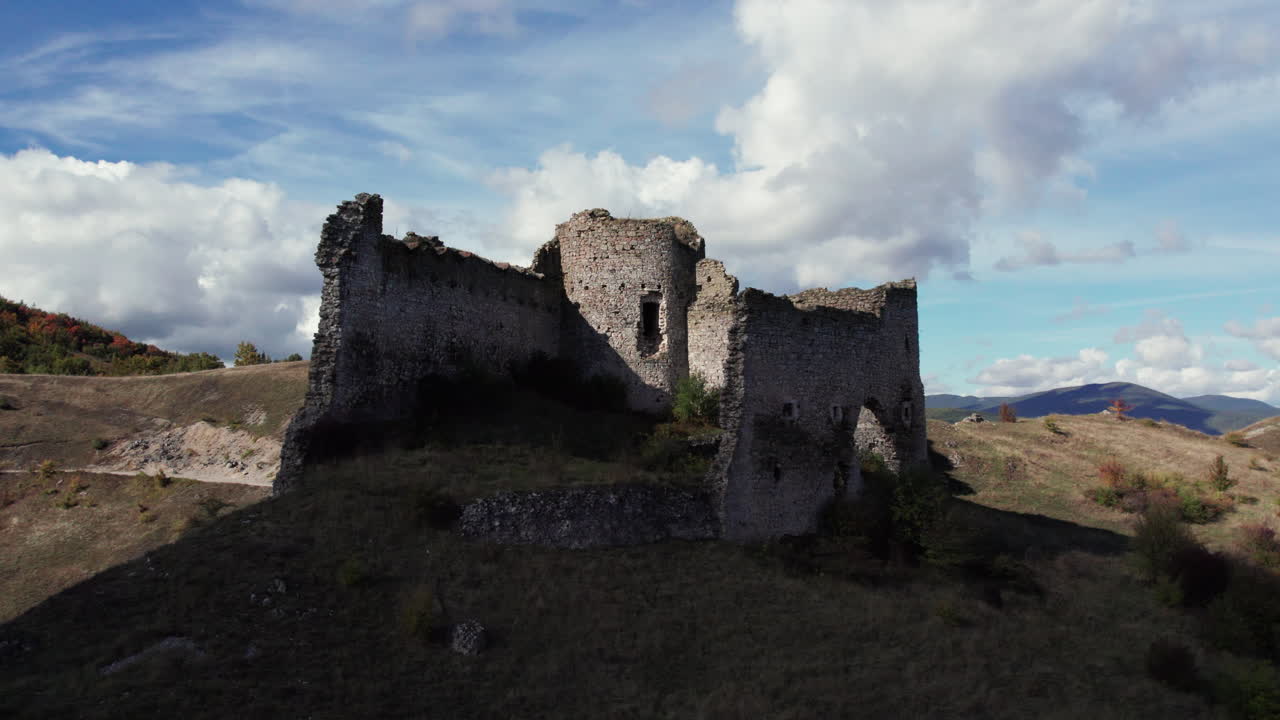 Aerial drone footage of the medieval Bjelaj Fortress ruins near Bosanski Petrovac, Bosnia and Herzegovina. The ancient stone walls rise above dry hills and scenic clouds, historic charm