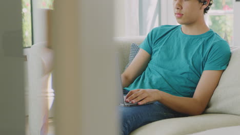 Adolescente-Usando-Auriculares-Inalámbricos-Sentado-En-Un-Sofá-En-Casa-Usando-Una-Computadora-Portátil