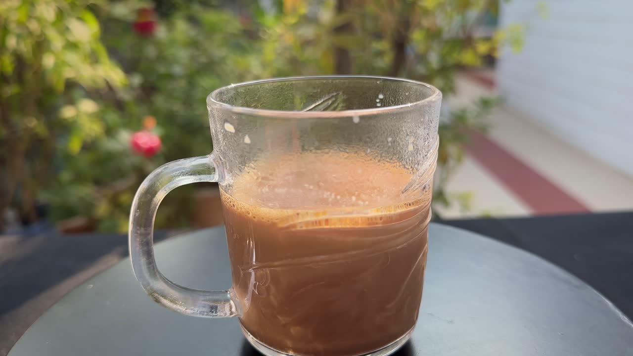 Primo piano di un giradischi con una tazza di tè caldo in vetro trasparente che ruota fluidamente su un piattino; vapore e schiuma visibili mentre la calda luce del giorno e un giardino sfocato creano un’atmosfera accogliente