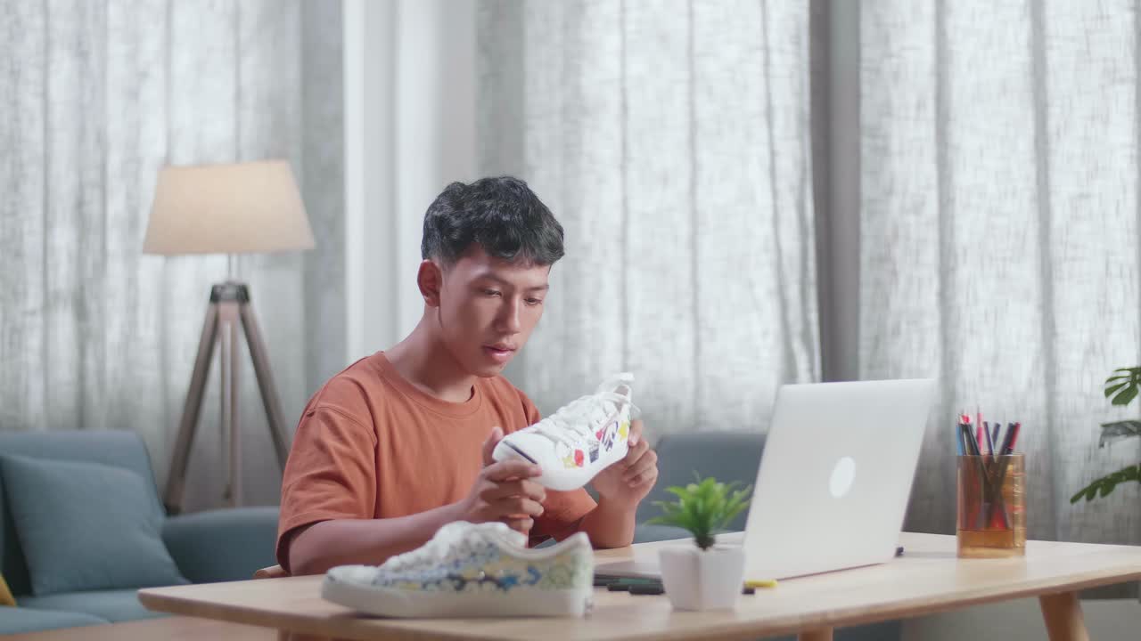 아시아 소년 신발 디자이너가 다채로운 패턴의 스니커를 들고 집에 있는 노트북의 사진과 비교합니다.