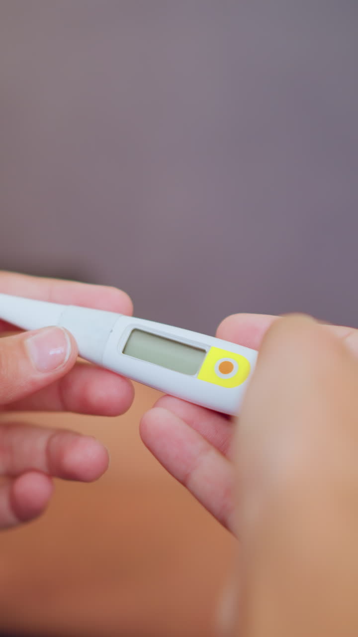 Close up of girl in maroon shirt raising hand while observing thermometer with calm serious expression as sick person lies blurred in background