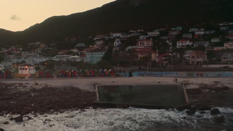 aerial drone shot captures ocean with tide pool and colorful beach shacks at sunset in greece. waves gently wash ashore, creating a serene scene.