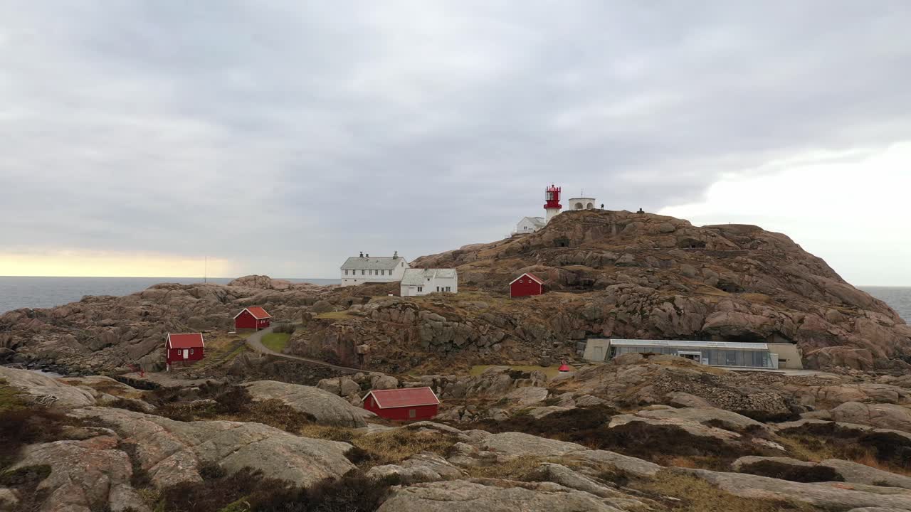 Lindesnes lighthouse forward moving low altitude aerial - Cliff foreground in lower frame with lighthouse tower and museum buildings in background against dramatic sky - Norway