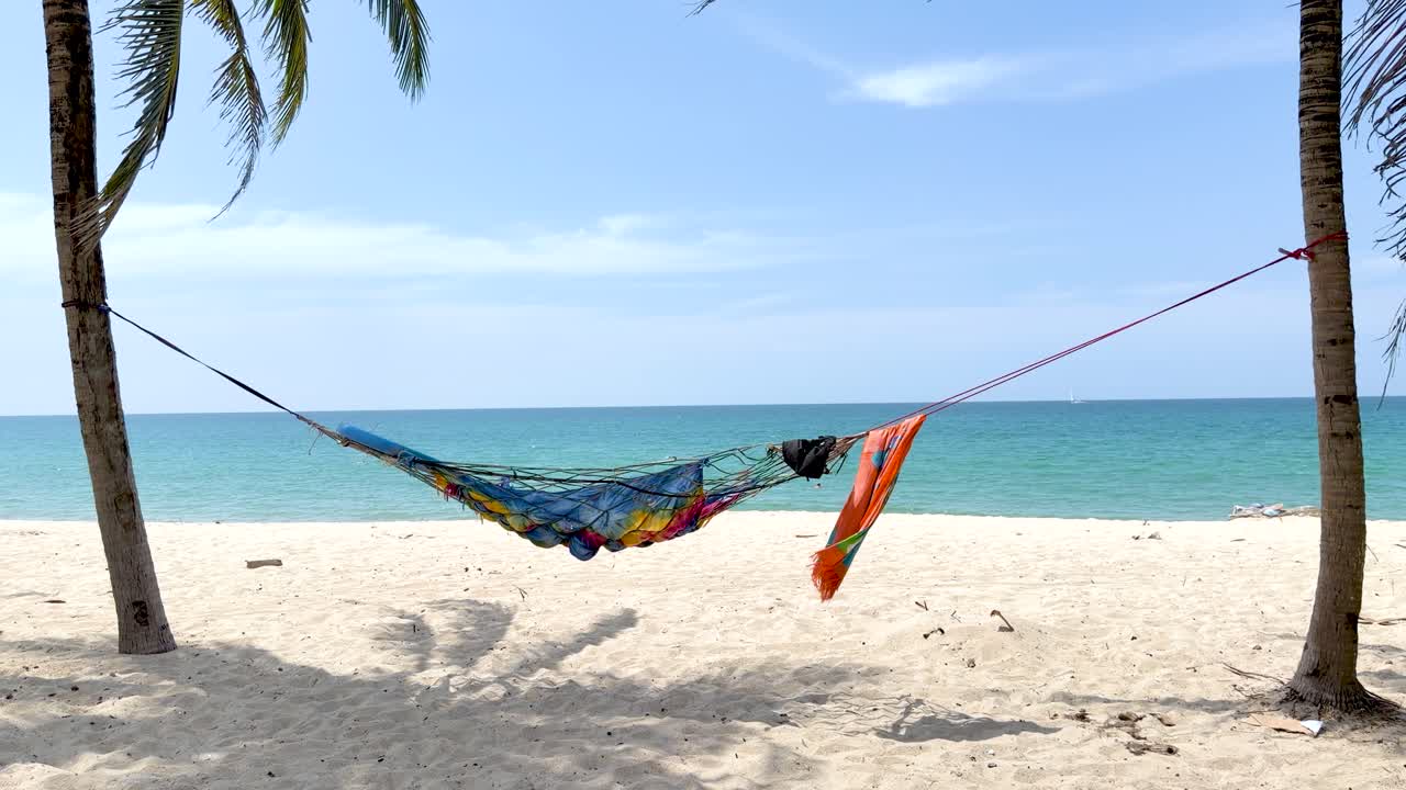Plano aberto estático de uma rede colorida balançando suavemente em uma ensolarada praia de areia branca.