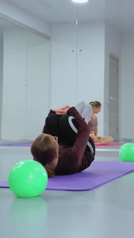 sesión de entrenamiento en grupo en un gimnasio bien iluminado con los participantes rodando hacia atrás en esteras, manos juntas en las rodillas, bajo el ojo atento de un instructor reflejado en un gran espejo de gimnasio